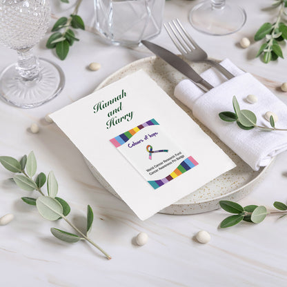 A personalized wedding place card for 'Hannah and Harry' styled on a speckled ceramic plate with a white napkin, silver fork and knife, and green olive branches on a light wood tabletop.